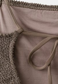 Hnedý texturovaný pletený odev s tenkými rebrovanými ramienkami, ktoré sú viazané cez očko na hladkom textilnom pozadí.