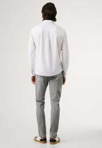 Mann steht mit dem Rücken zur Kamera, trägt ein weißes Langarmshirt, graue Jeans und weiße Sneakers mit roten und blauen Akzenten.