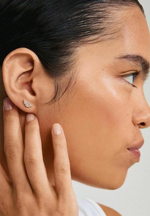 Boucle d'oreille en ton or avec des strass clairs, présentant un design en forme de vague. Gros plan sur l'oreille et la main d'un modèle. Texture de peau lisse.