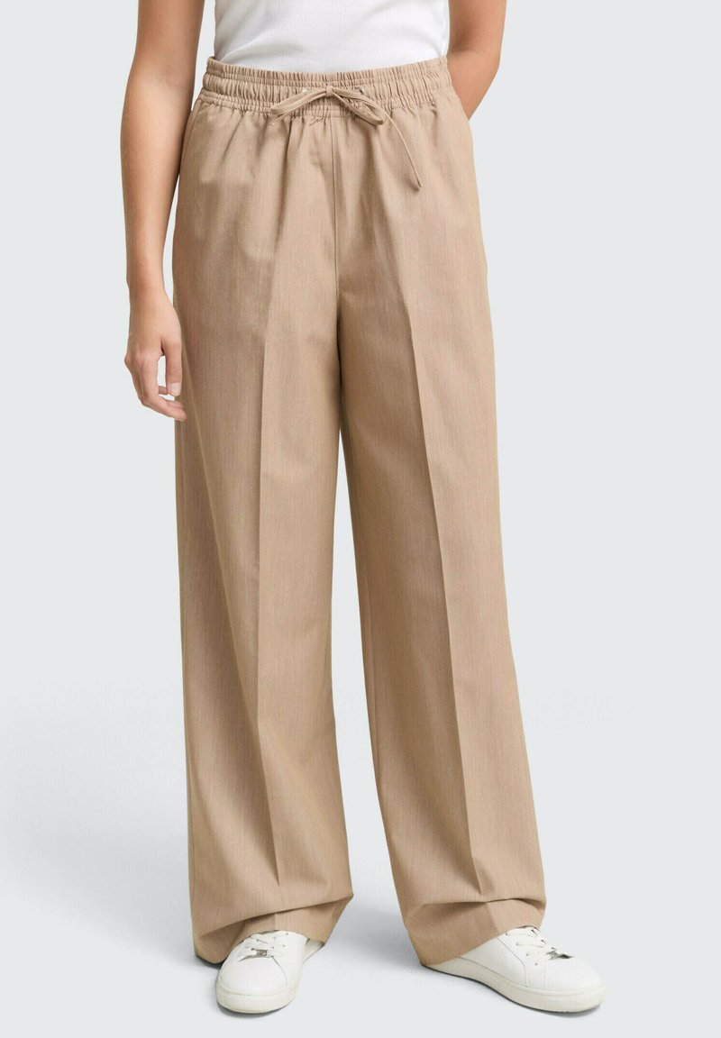 Pantaloni beige a gamba larga con pieghe, fascia elastica in vita e chiusura con cordino. Indossati con sneakers bianche su uno sfondo grigio chiaro.