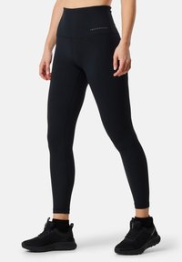 Leggings noirs taille haute en matériau élastique, avec une texture lisse et le logo de la marque "SWEDMOUNT" sur la taille.