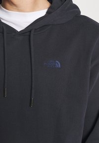 Námořnicky modrá mikina s kapucí, vybavená přední kapsou, nastavitelným tažným provázkem a malým vyšitým logem na levé straně hrudi. Měkká tkanina s žebrovanou strukturou.