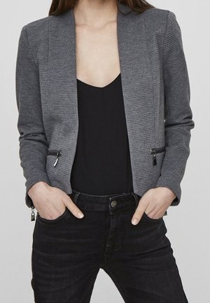 Mujer que lleva un blazer gris entallado de canalé con bolsillos con cremallera sobre una camiseta negra y jeans negros con las manos en los bolsillos.