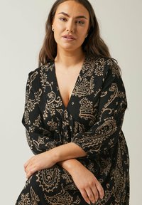 Schwarzes Kleid mit beigem Paisleymuster. Hat einen tiefen V-Ausschnitt und dreiviertel Länge Ärmeln. Texturiertes Material sorgt für visuelles Interesse.