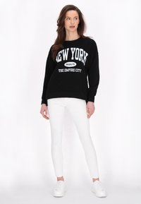 Nő áll, rajta fekete kapucnis pulóver, amelyen a "New York Brooklyn The Empire City" felirat olvasható, fehér nadrágban és fehér sportcipőben, fehér háttér előtt.