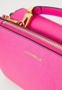 Pink textured leather handbag with a gold zipper and hardware, featuring a logo in gold. Compact shape with a short handle.