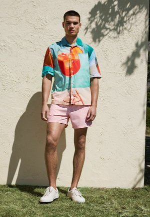 Young man standing outdoors against beige wall wearing colorful short-sleeve shirt with large drink print, pink striped shorts, and white sneakers.