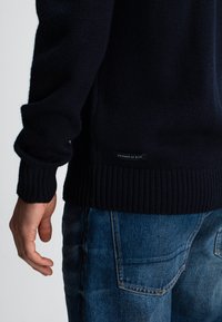 Man in een marineblauwe gebreide trui met een "Butcher of Blue" label en blauwe spijkerbroek, met een hand ontspannen naast zich.