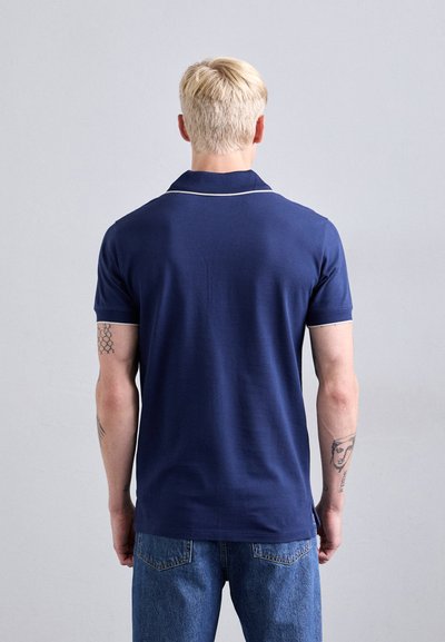 Tricou polo albastru marin cu guler ribat și piping alb contrastant, fabricat din bumbac moale, purtat cu blugi albastru deschis, vedere din spate.