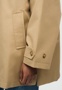 Κοντινό πλάνο μανικιού μπεζ παλτού με κουμπωτό καπάκι μανσετών και ορθογώνια πλαϊνή τσέπη, που φοριέται πάνω από μπλε τζιν, δείχνοντας μια χαλαρή θέση του χεριού.