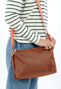 Person med blå jeans og en hvid langærmet skjorte med grønne striber, der holder en struktureret rustfarvet crossbody-taske med en læderrem.