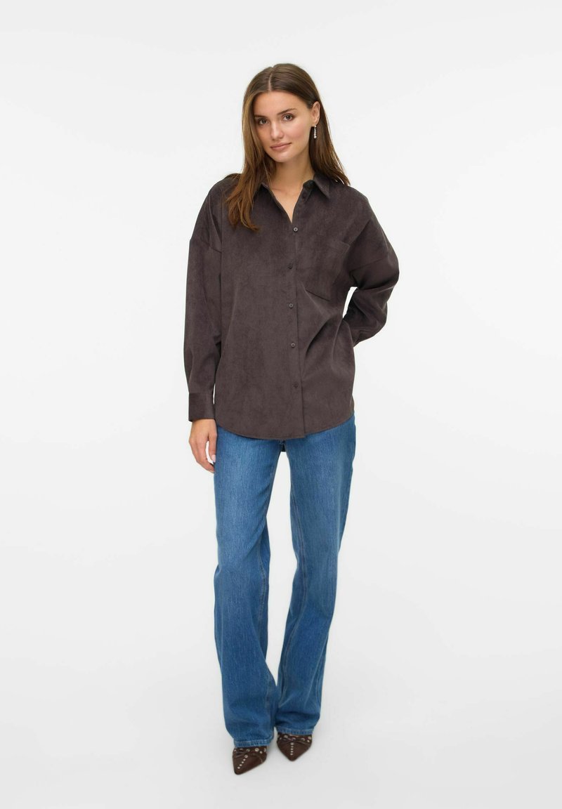 Donkerbruine suède button-up shirt met een relaxte pasvorm, enkele borstzak, opgerolde mouwen, gecombineerd met lichtblauwe flared jeans en puntige schoenen.