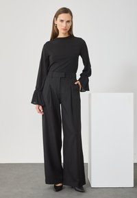Maglietta nera a maniche lunghe con collo alto e maniche a campana, abbinata a pantaloni neri a vita alta e gamba larga. Tessuto morbido, design minimalista.