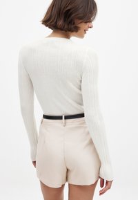 Ribbed valkoinen neule pitkähihaisilla hihoilla, yhdistettynä vaaleanbeigeihin mittatilaustyönä tehtyihin shortseihin ja mustaan vyöhön, joka esittelee tiukkaa siluettia takaa.