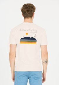 Ljusbeige t-shirt med en grafisk design av en sol, berg och palmträd i mörkblå, grön, gul och orangea ränder på baksidan.