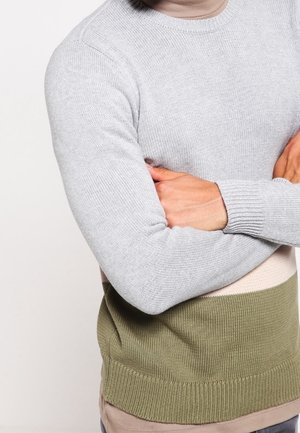 Ein Mann verschränkt die Arme und trägt einen Pullover im Farbblockstil mit hellem Grau oben und Olivgrün unten, stehend vor einem schlichten hellen Hintergrund.