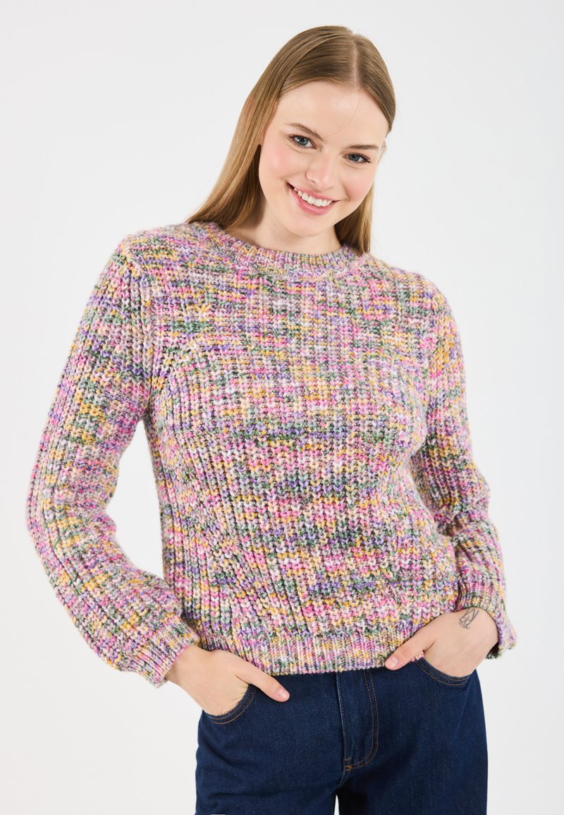 Jeune femme souriante, se tenant avec les mains dans les poches d'un jean bleu foncé, portant un pull en tricot multicolore devant un fond blanc uni.