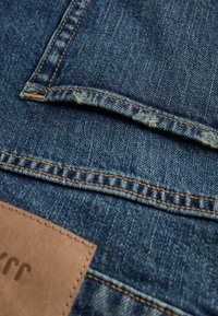 Primer plano de unos vaqueros azules de mezclilla que muestran un borde deshilachado en un bolsillo trasero y una etiqueta de marca de cuero marrón claro.