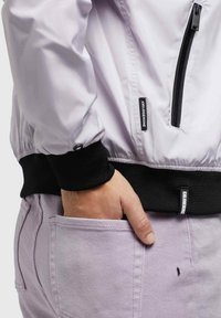 Personne portant un pantalon violet clair avec la main dans la poche arrière et une veste blanche avec fermeture éclair noire et poignets noirs, étiquettes de marque visibles sur la veste.