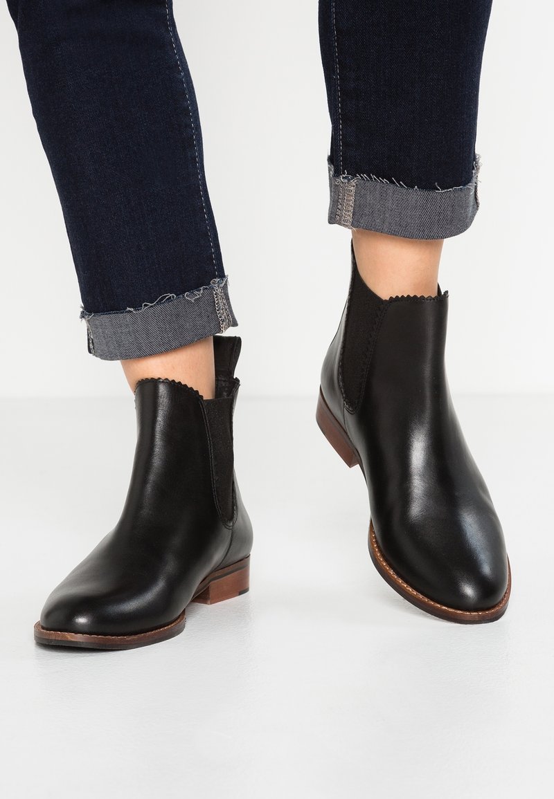 Personne portant des bottines en cuir noir aux bords festonnés et un jean foncé retroussé, debout sur un sol blanc.