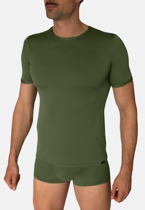 Mand iført en tætsiddende olivengrøn kortærmet skjorte og matchende boxershorts, stående mod en ensfarvet baggrund.