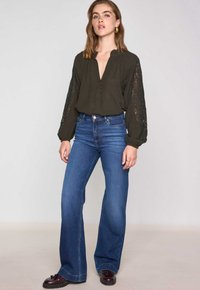 Blusa verde scuro con bottoni e maniche in pizzo, abbinata a jeans di denim a campana. Le scarpe presentano una finitura lucida scura.