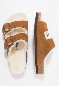 Bruna mockaskinnsandaler med två justerbara remmar och pälsfoder, med en texturerad mörkbrun sula och en glittrande detalj på kanten.