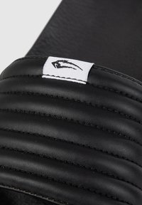 Sandal in gomma nera con superiore trapuntato e scanalature orizzontali. Un'etichetta bianca con un logo è visibile sulla cinghia.