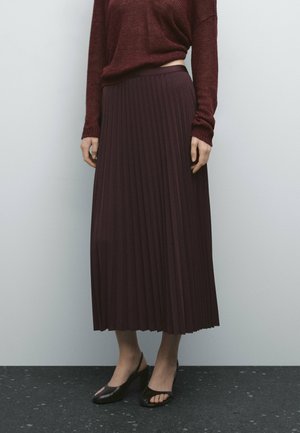 Person iført en mørk plisseret midi-nederdel, matchende strikket sweater med lange ærmer og sorte åbne tå-hæle, stående på et sort gulv mod en lys væg.