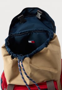 Rucsac roșu și bej cu închidere tip rolă, având un interior bleumarin, șnur de strângere și o patch cu logo în interior. Textură durabilă a țesăturii.