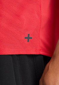 Camiseta deportiva roja con un patrón texturizado y detalle de logo negro en forma de "+", combinada con pantalones cortos negros que presentan una costura visible.