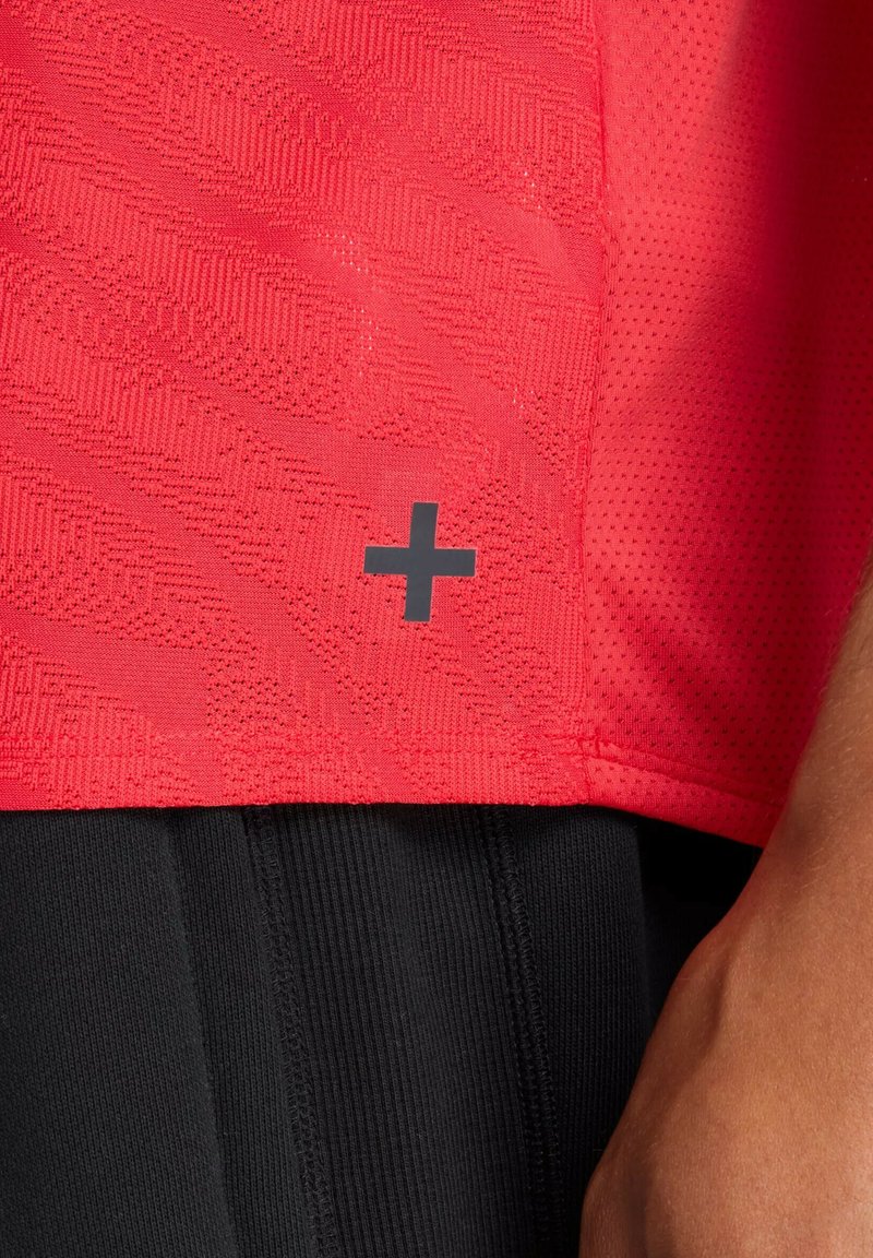 Rotes Sportshirt mit strukturiertem Muster und schwarzem "+"-Logo, kombiniert mit schwarzen Shorts mit sichtbarer Naht.