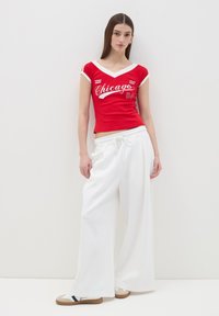 Camiseta roja con escote en V y gráfico "Chicago 04", combinada con pantalones anchos blancos. La modelo lleva zapatillas beige con un acento azul marino.