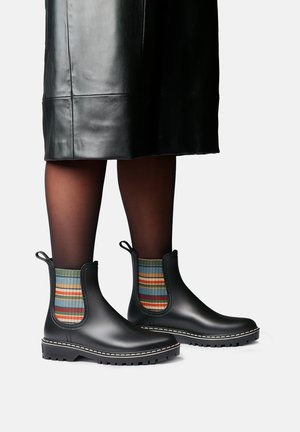 Bottines en cuir noir avec des panneaux latéraux élastiques rayés multicolores, portées avec des collants noirs transparents et une jupe en cuir noire.