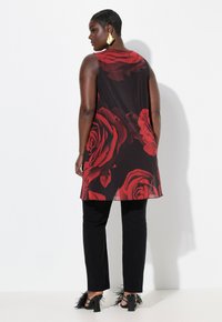 Tunic nera con grande stampa di rose rosse, indossata sopra pantaloni neri, con piume su tacchi neri. La schiena della modella è rivolta verso la macchina fotografica.