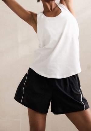 Žena nosící bílý nátělník bez rukávů a černé sportovní šortky, natahující paže na neutrálním pozadí.