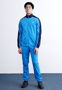 Μπλε αθλητική φόρμα με ζακέτα με φερμουάρ, ναυτικές λεπτομέρειες και αντίστοιχες παντελόνες. Φοριέται με μαύρα παπούτσια, διατυπωμένο σε απλό φόντο.