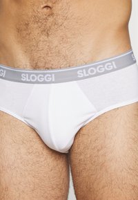 Bela bombažna spodnjica z sivim elastiko v pasu, na katerem je v belih črkah napis "SLOGGI". Klasičen kroj, gladka tekstura.