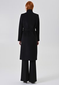 Zwarte wollen jas met hoge kraag, een ceintuur in de taille en lange mouwen. Gladde textuur en ontwerp tot de knie. Uitlopende zwarte broek zichtbaar eronder.