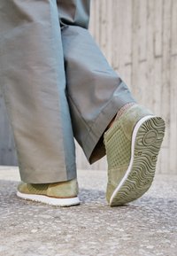 Žali od suede medvilniniai sportbačiai su tinklelio akcentais, balta guma padanga ir tekstūruotas dugnas, derinami su šviesiais pilkais plačiais kelnių.