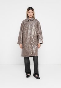 Leopard print, shiny vinyl coat with a collared neckline, button front, and loose fit, paired with dark, wide-leg pants and flat shoes.
