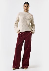 Dikke romige gebreide trui met ribbelstructuur en drie knopen bij de schouder, gecombineerd met wijde burgundy corduroy broeken.