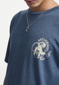 Camiseta de algodón azul marino con un gráfico en blanco de un astronauta que dice "ANTI-GRAVITY BOTTLE SHOP". Cuello redondo y corte relajado. Cadena de plata visible.
