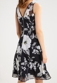 Svart blommig klänning med en genomskinlig överdel, V-ringning och en dragkedja bak. Stora vita blommor med gröna accenter på en svart bakgrund.