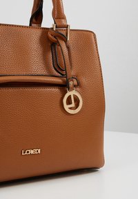 Sac à main marron texturé avec un logo doré et un porte-clés en accent, doté d'une poche zippée à l'avant et d'un bord contrastant noir.