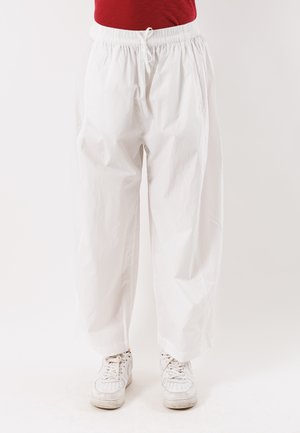 Pantaloni bianchi larghi con coulisse indossati con sneakers bianche e top rosso, mostrati dalla vita ai piedi su uno sfondo semplice.