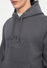 Sudadera con capucha de color gris oscuro con un logotipo texturizado bordado "ORIGINALS Studio" en el pecho. Cuenta con cordones y un bolsillo tipo canguro.