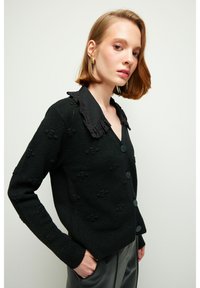 Svart stickad cardigan med ett texturerat mönster, en svart spetskrage och framknappar. Ärmarna är långa med ribbade manschetter.