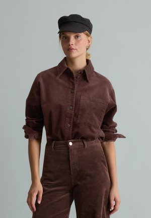Jeune femme portant une chemise et un pantalon en velours côtelé marron foncé avec les manches retroussées et une casquette noire, debout devant un fond gris uni.