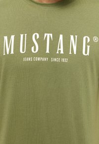Olíva zöld pamut póló, amelyen nagy fehér betűkkel a "MUSTANG" felirat szerepel, alatta kisebb betűkkel a "JEANS COMPANY SINCE 1932" felirattal.